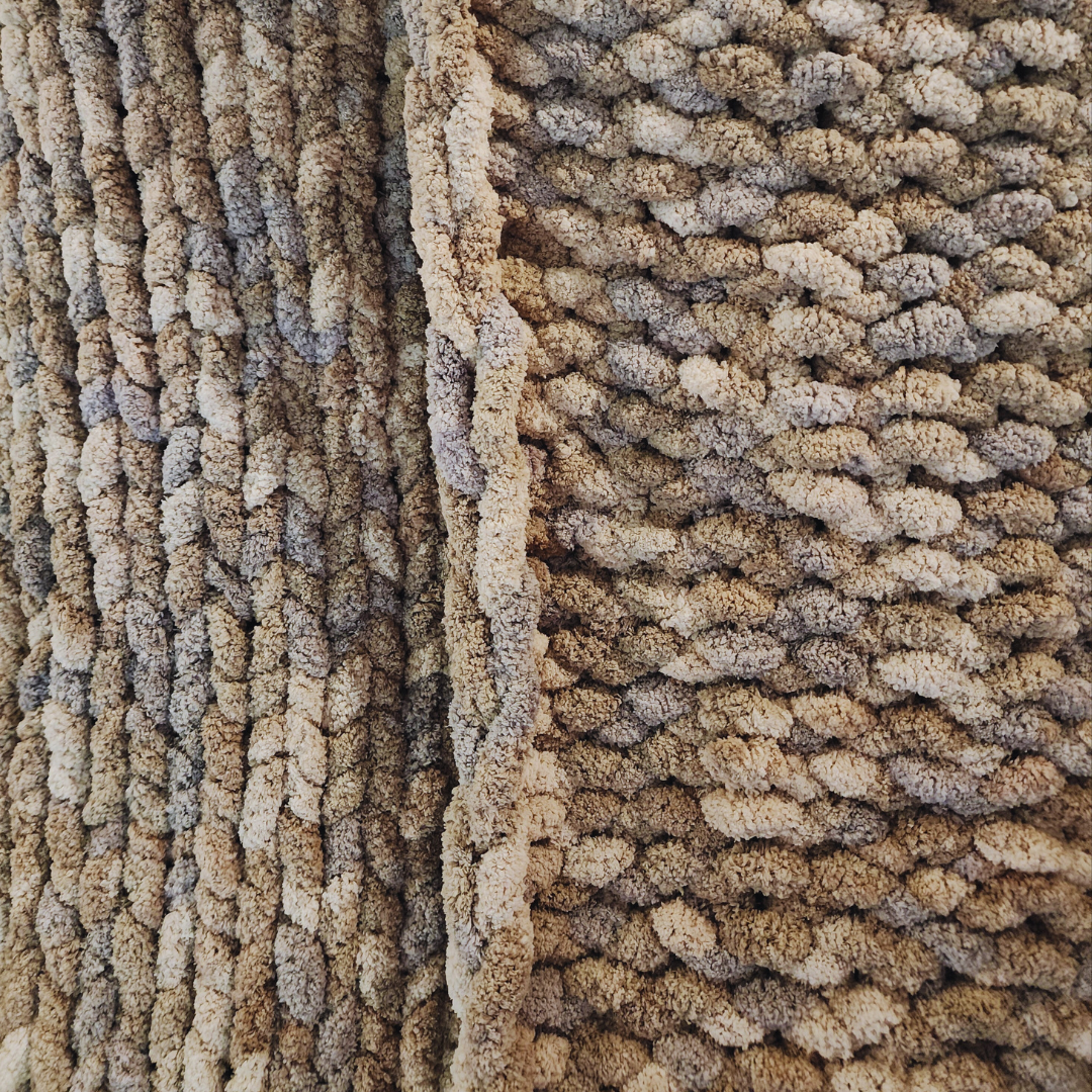 A close-up overhead view of a handmade chunky knit blanket, showing its soft, textured surface and multicolor pattern in shades of gray, white, and light brown, with a vertical fold.
