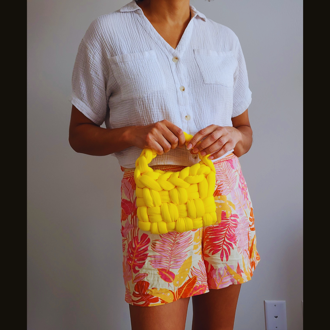 A person wearing a white shirt and tropical print shorts holds a small handmade yellow basket weave handbag by its braided handle, posed against a white wall.