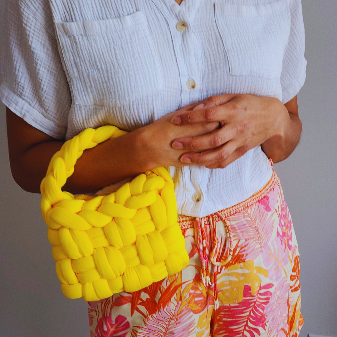 A close-up of a person's hands resting on a small handmade yellow basket weave handbag, showing the bag's textured surface and braided handle, against a background of tropical print shorts.