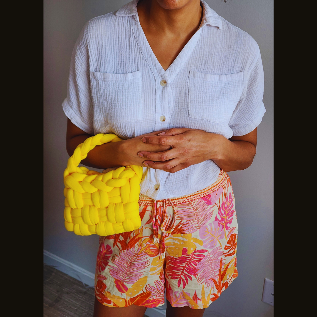 A person wearing a white shirt and tropical print shorts stands holding a small handmade yellow basket weave handbag with both hands clasped over it.