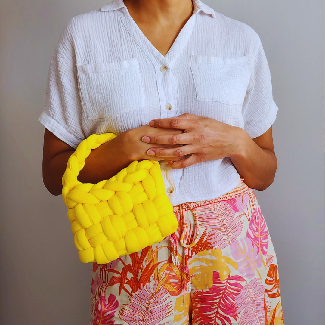 A person wearing a white shirt and tropical print shorts holds a small handmade yellow basket weave handbag close to their body with both hands, showing the bag's textured surface.