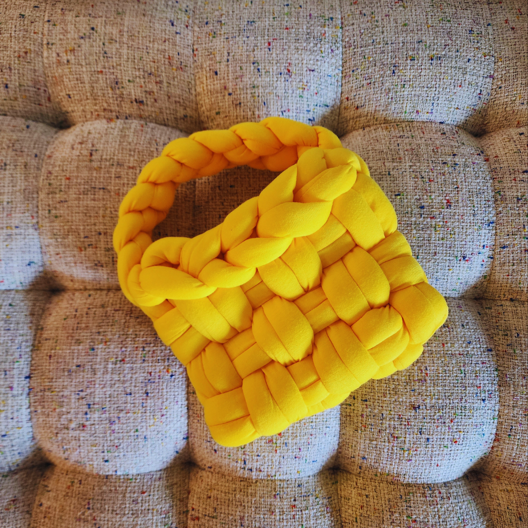 A small handmade basket weave handbag in vibrant yellow, with a thick braided handle, lying flat on a multicolored speckled fabric surface.
