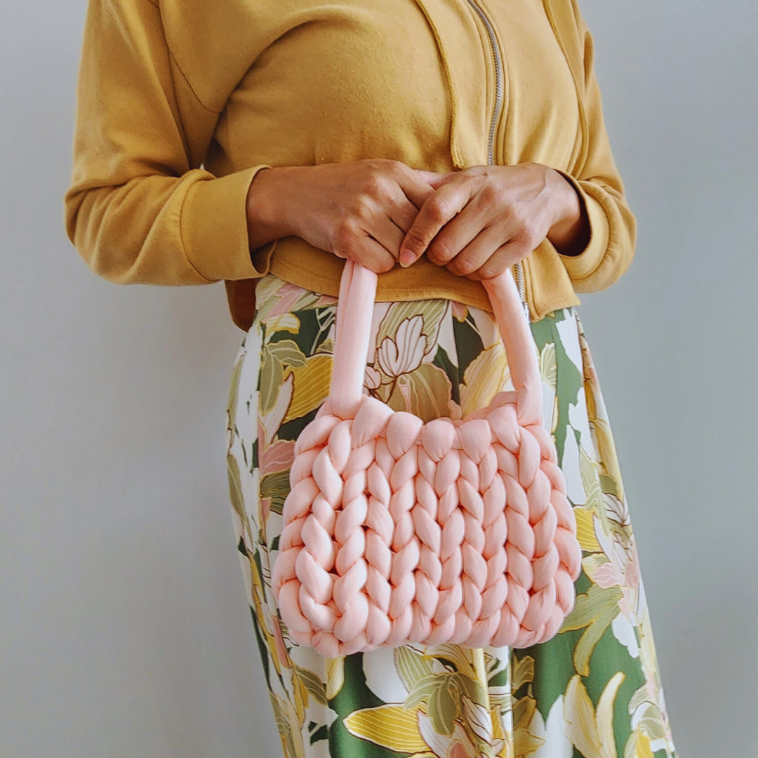 A person wearing a yellow hoodie and a floral skirt holds a small handmade single stitch knit bag in light coral pink with both hands, positioned in front of their torso.