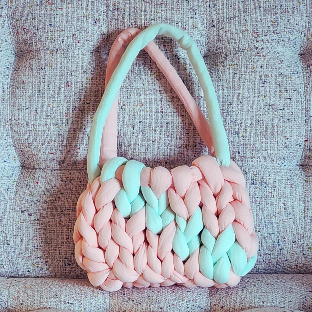 A small handmade single stitch knit bag in light coral pink with two vertical stripes of mint green, and two-toned handles, sitting upright on a textured pink and white upholstered surface.