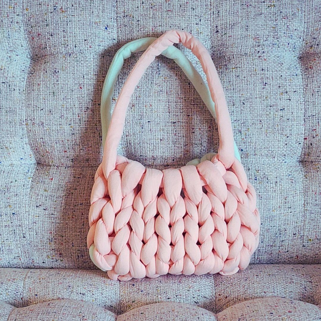 A small handmade single stitch knit bag in light coral pink with a hint of mint green on the handles and sides, sitting upright on a textured pink and white upholstered surface.