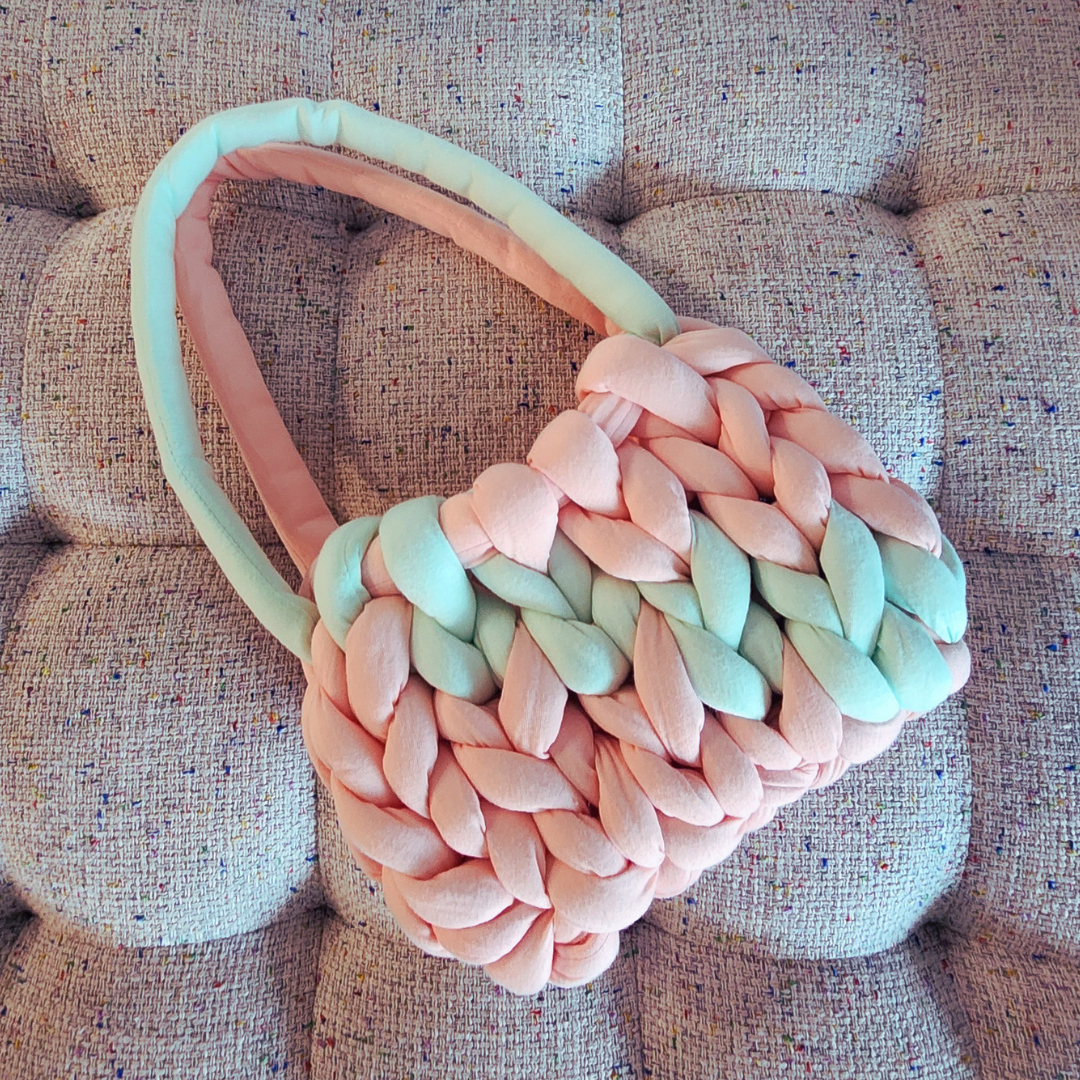 A small handmade single stitch knit bag in light coral pink with a broad diagonal stripe of mint green, and two-toned handles, positioned diagonally on a textured pink and white upholstered surface.