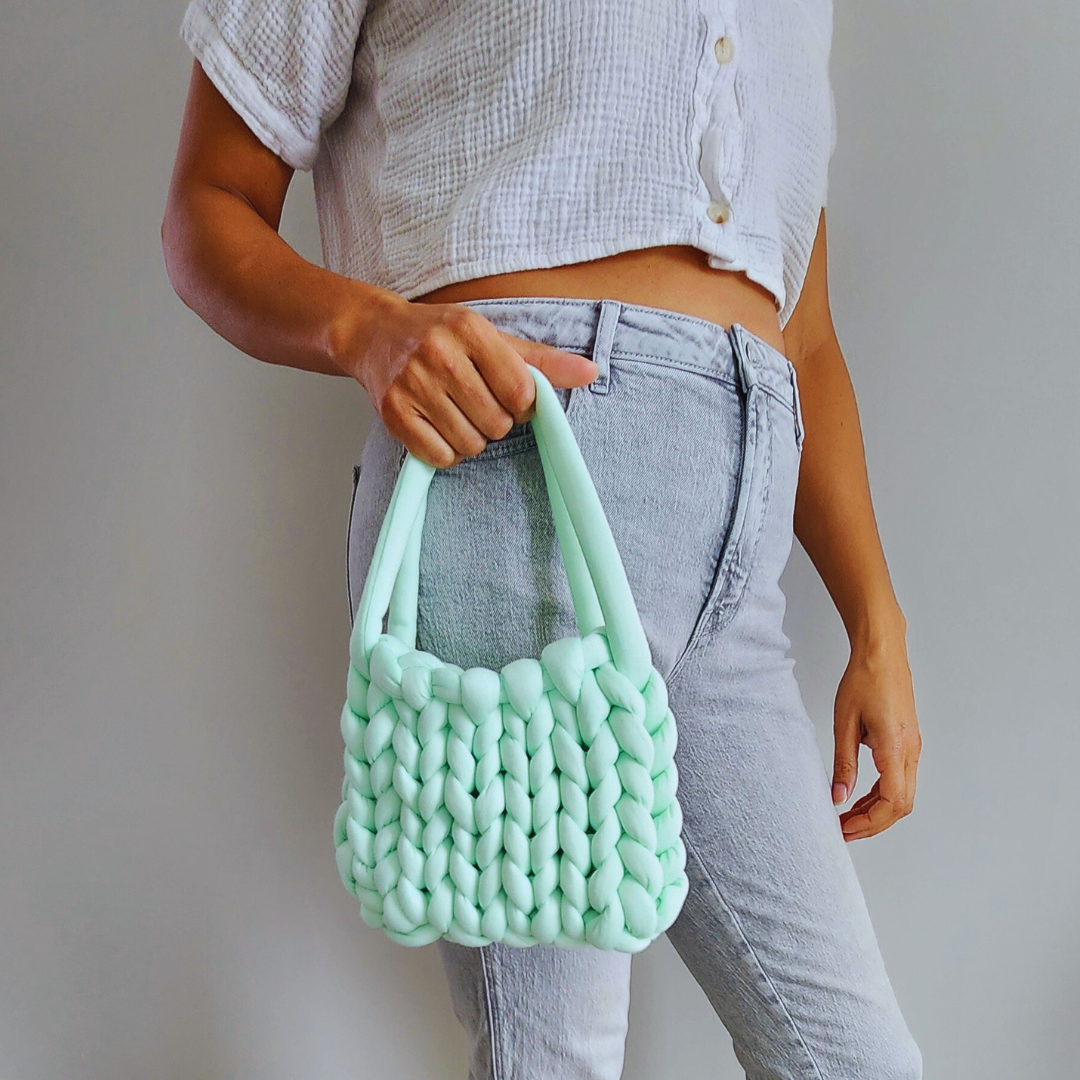 A person wearing a white cropped shirt and light gray jeans holds a small handmade single stitch knit bag in light mint green by its double straps, posed against a light gray wall.