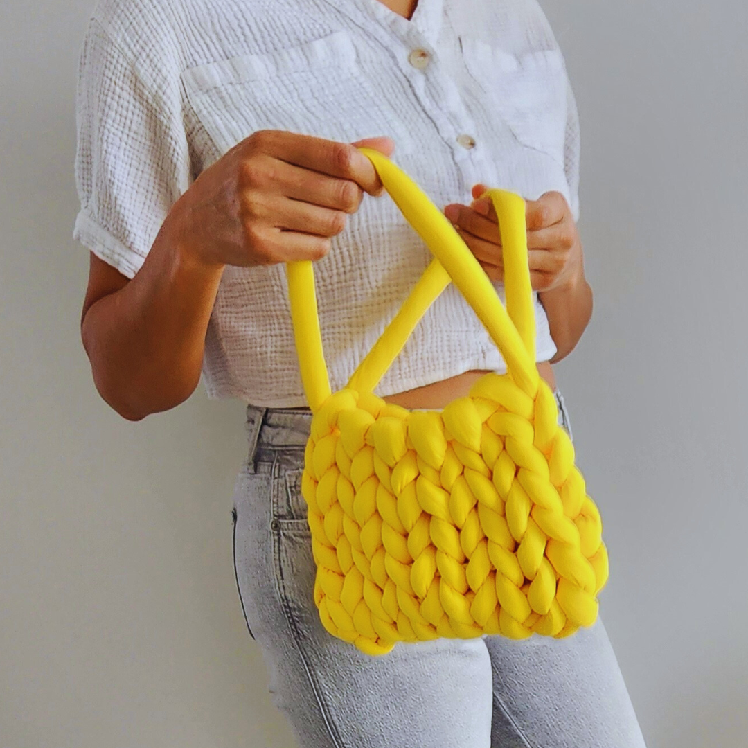 A person wearing a white cropped shirt and light gray jeans holds a small handmade single stitch knit bag in bright yellow with both hands, showing the bag's woven texture, against a light gray wall.