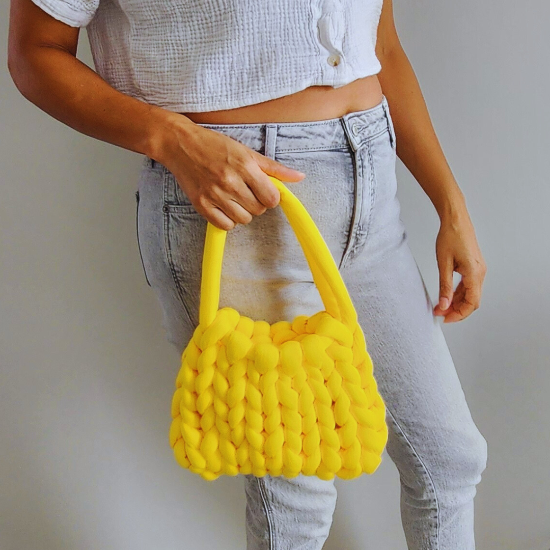 A person wearing a white cropped shirt and light gray jeans holds a small handmade single stitch knit bag in bright yellow by its double straps, posed against a light gray wall.