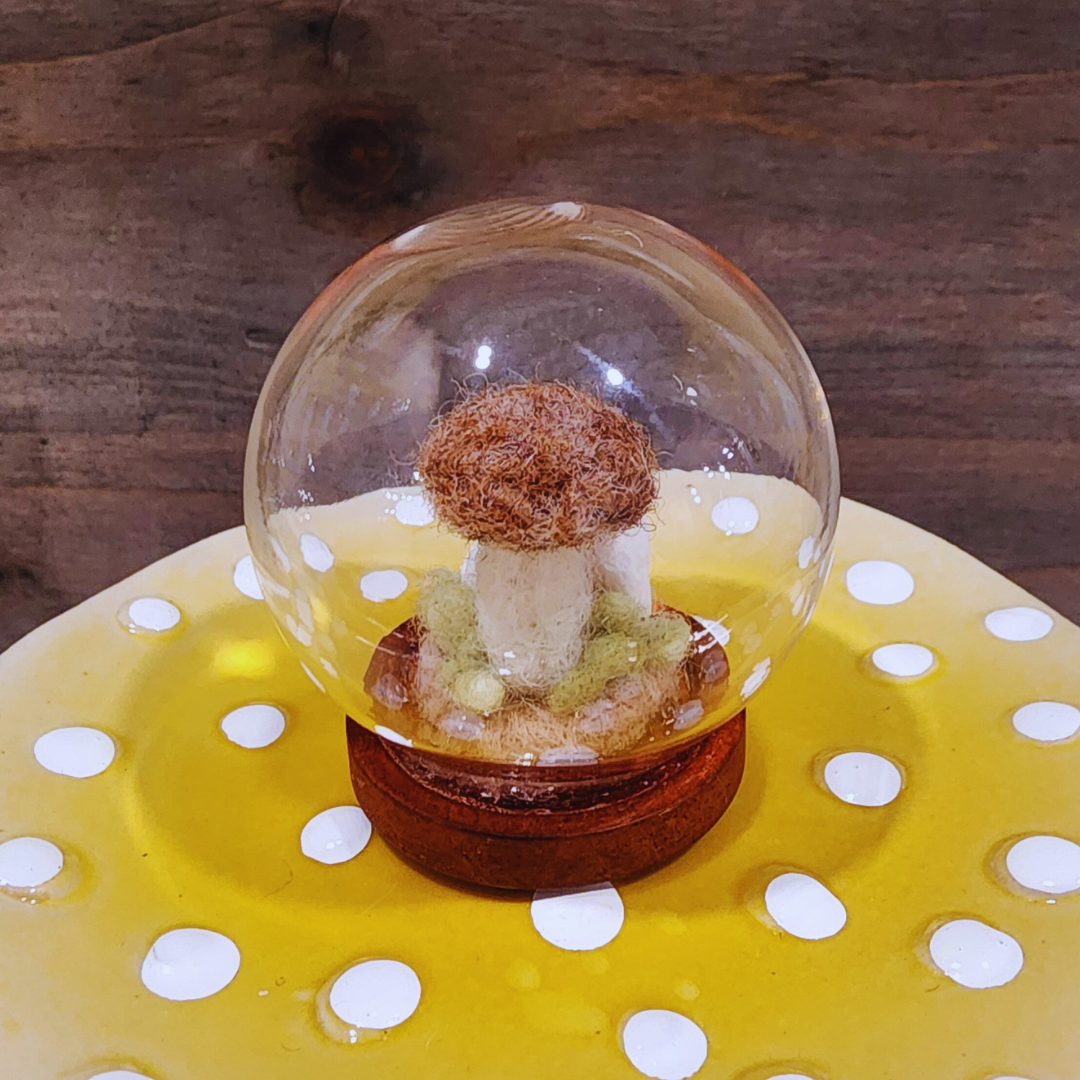 A handmade felted Brown Cap mushroom terrarium in a glass globe, featuring green moss on a dark wooden base.
