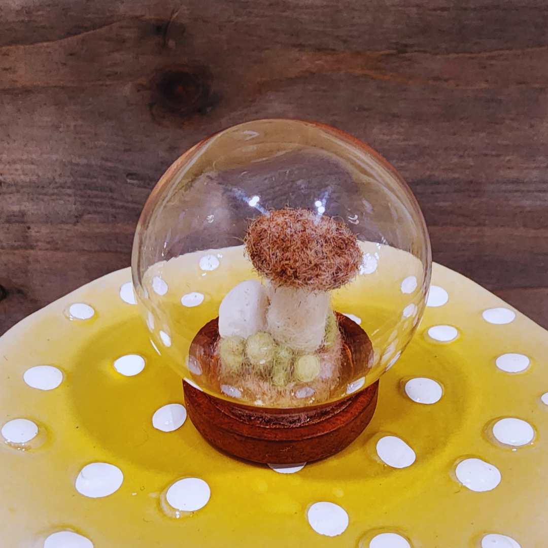 A handmade felted Brown Cap mushroom terrarium in a glass globe, featuring white felt stones beneath the cap.