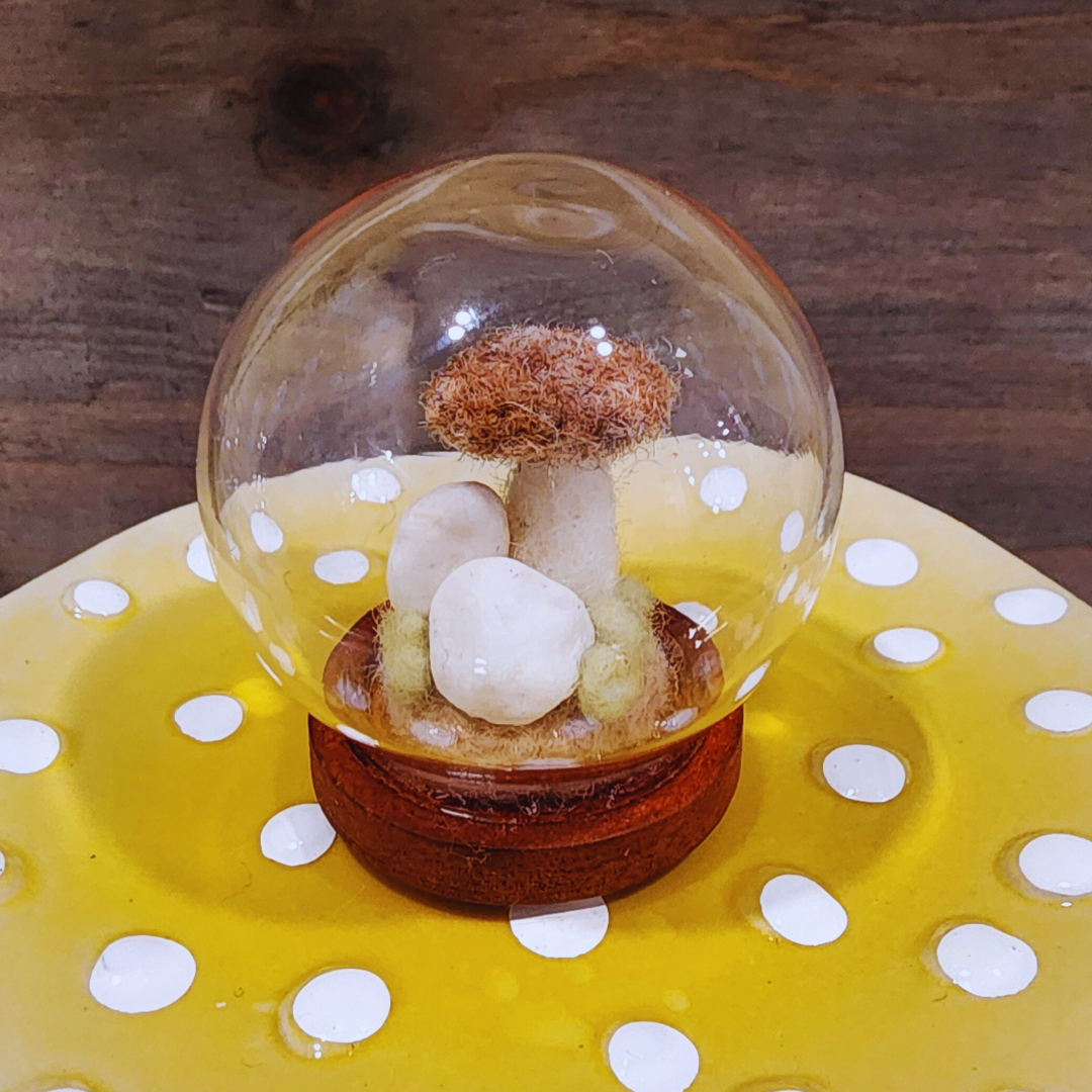A handmade felted Brown Cap mushroom terrarium in a glass globe, featuring white felt stones and a dark wooden base.