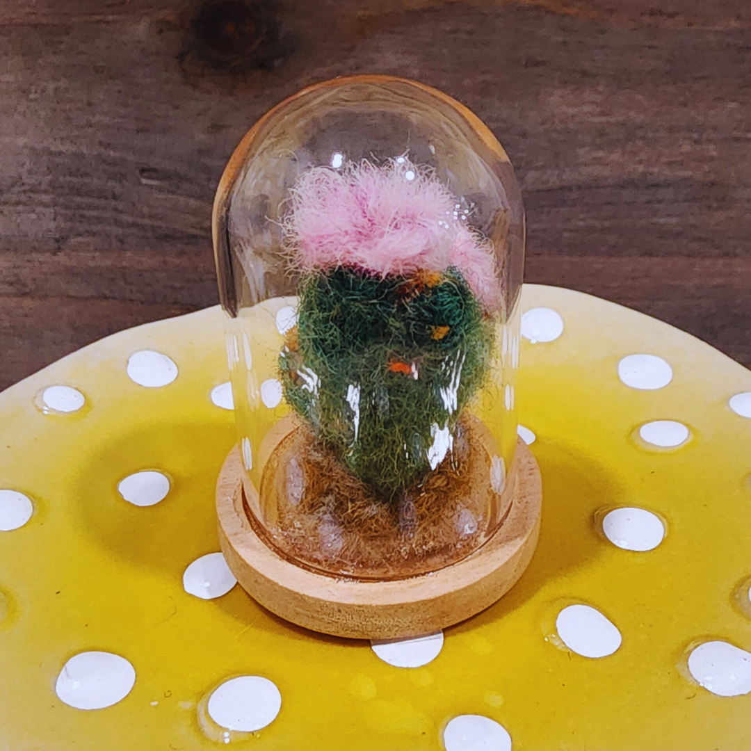 A close-up of a handmade felted Flowering Cactus terrarium with a large pink bloom in a glass dome.