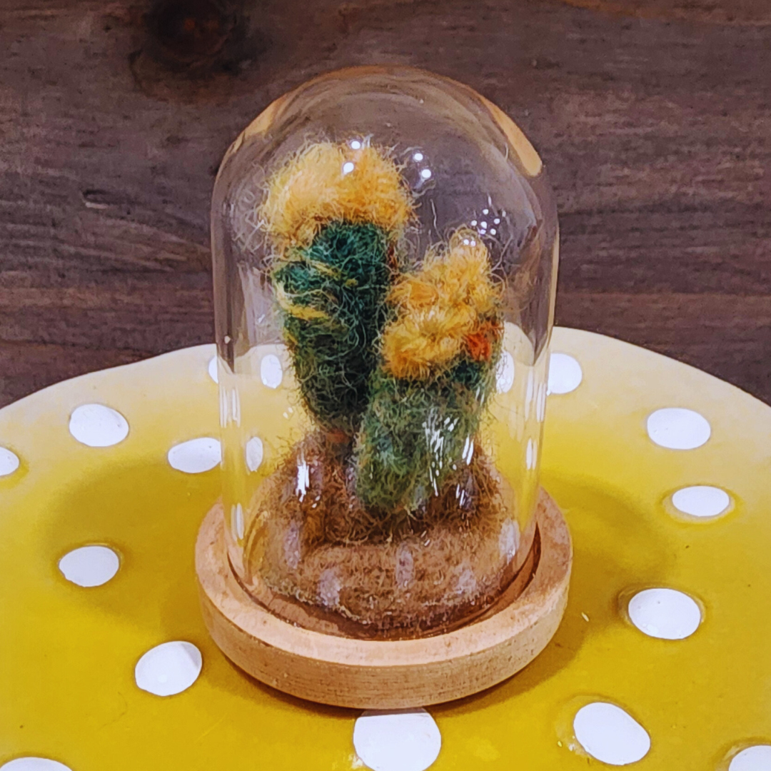 A close-up of a handmade felted Flowering Cactus terrarium with yellow blooms in a glass dome.
