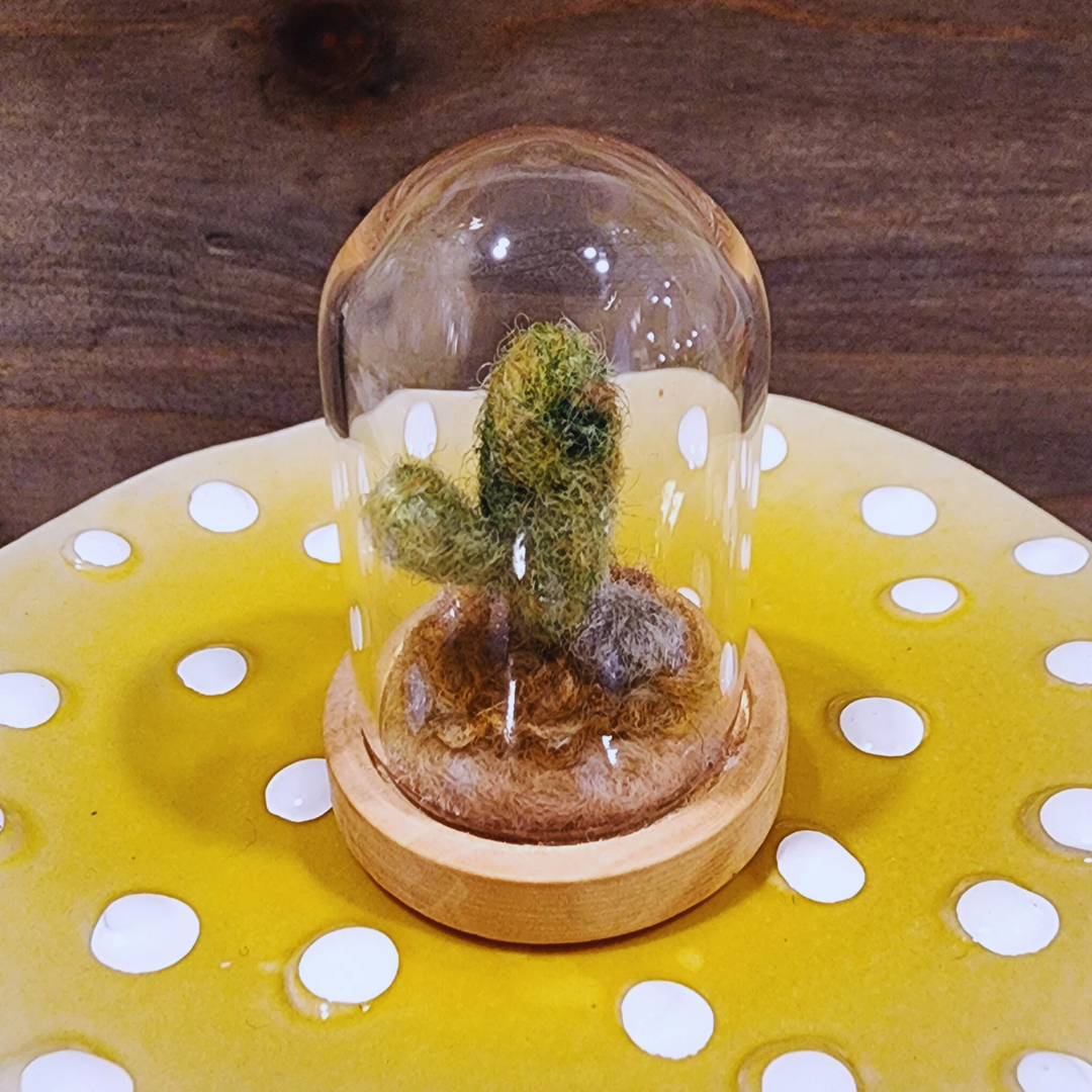 A handmade needle-felted Saguaro cactus terrarium in a glass dome, sitting on a light wood base.