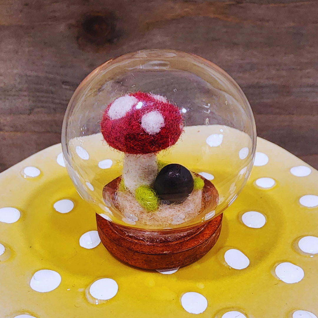 A handmade needle-felted red and white spotted Toadstool mushroom terrarium in a glass globe on a wooden base.