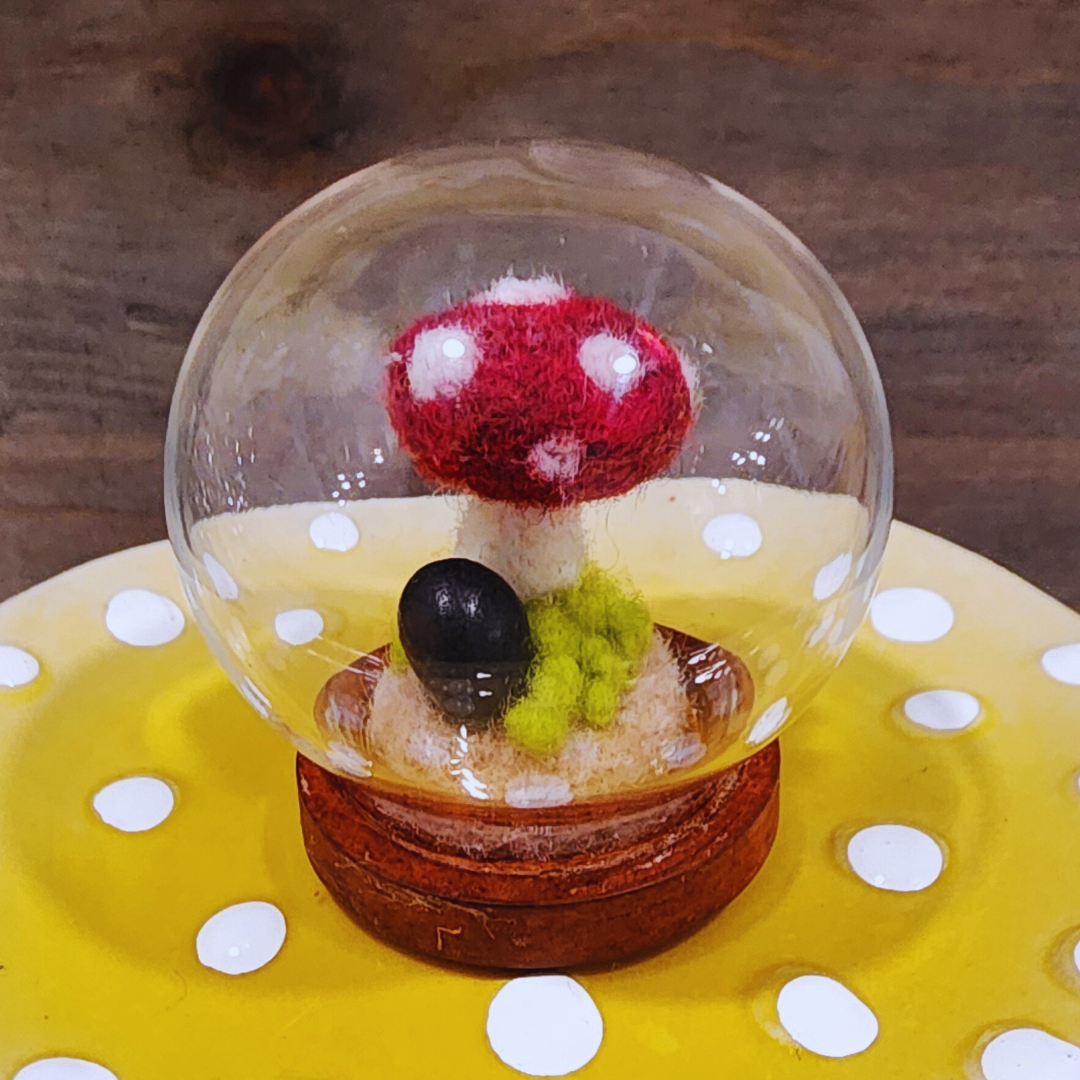 A handmade felted red and white Toadstool mushroom terrarium in a glass globe, showing green moss and a black stone.