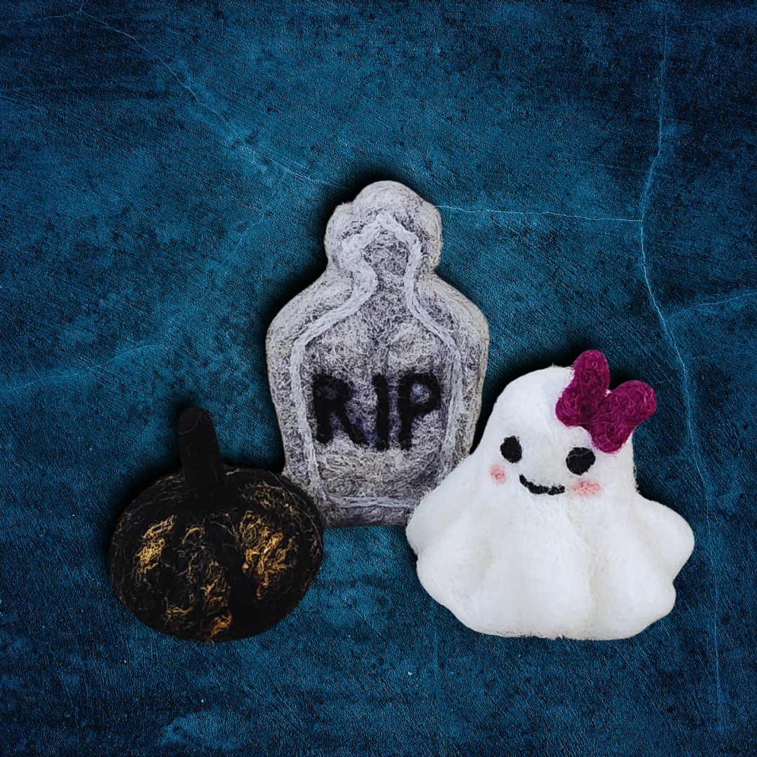 A group of handmade felted Halloween figurines, including a gray tombstone with "RIP," a black pumpkin with a black stem, and a white ghost with a purple bow, arranged on a textured blue surface.