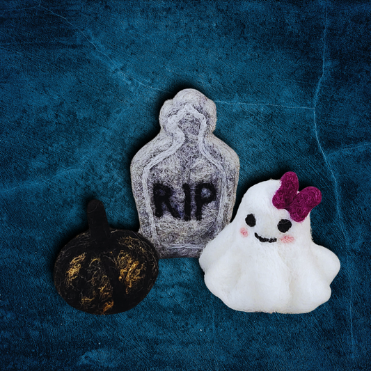 A group of handmade felted Halloween figurines, including a gray tombstone with "RIP," a black pumpkin with a black stem, and a white ghost with a purple bow, arranged on a textured blue surface.