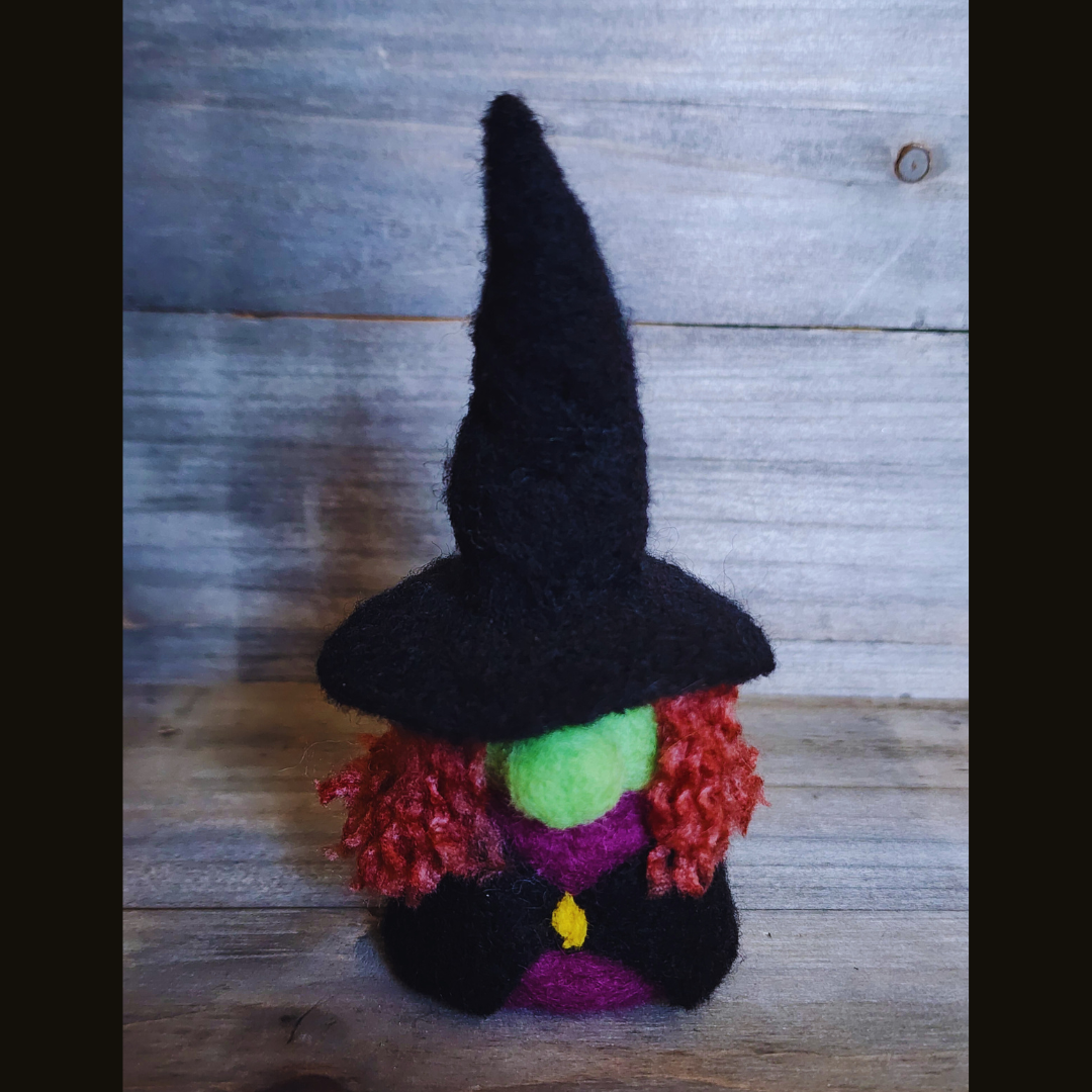 A handmade needle-felted witch figurine with a black pointy hat, a bright green face, a purple body, a yellow buckle, and curly orange-red hair, standing centered against a wooden background.