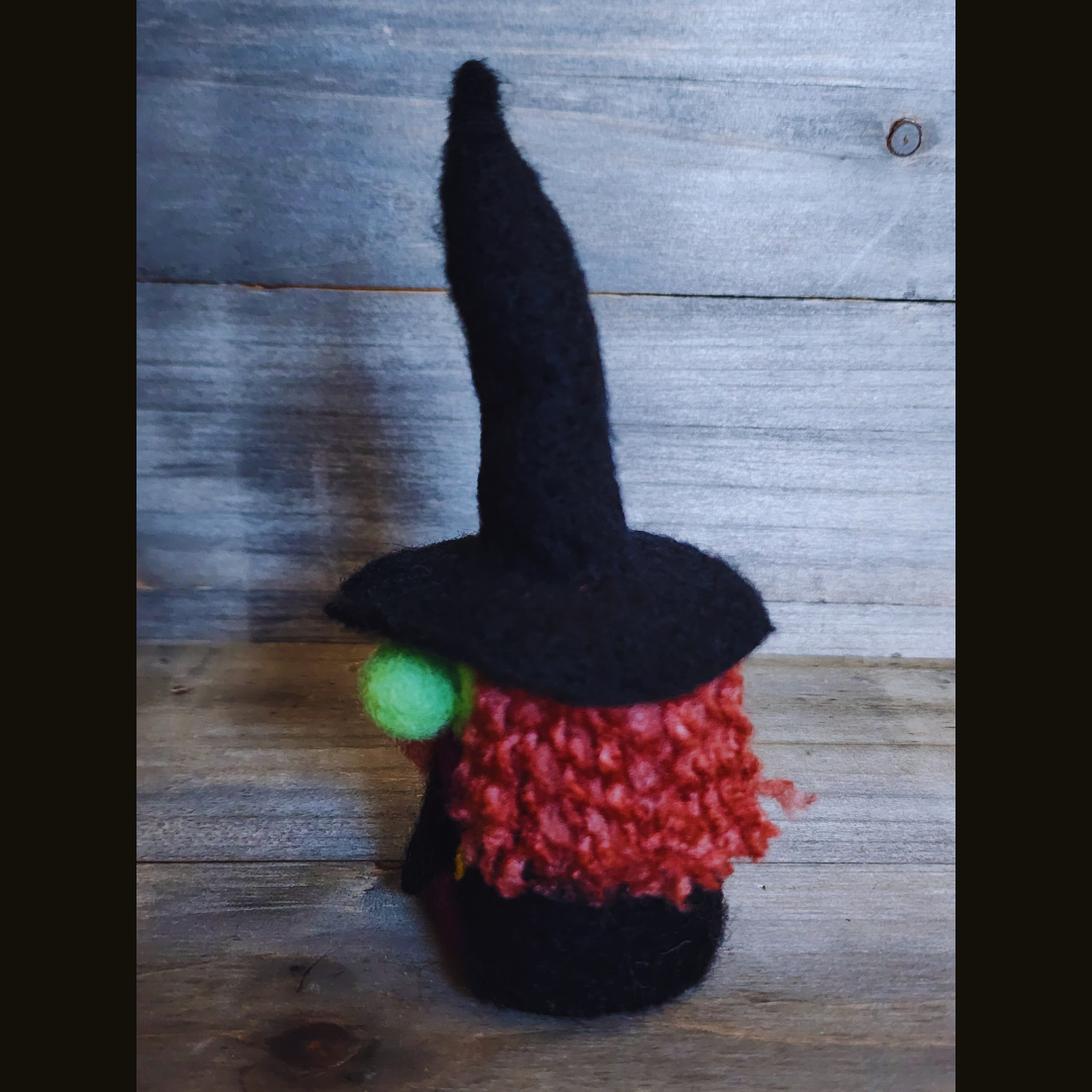 A handmade needle-felted witch figurine with a black pointy hat, a bright green face, a purple body, and curly orange-red hair, viewed from a side angle against a wooden background.