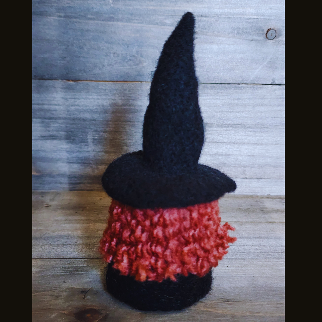 A handmade needle-felted witch figurine with a black pointy hat and curly orange-red hair, viewed from a slightly angled back position against a wooden background.