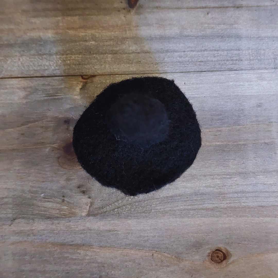 An overhead view of a handmade needle-felted witch figurine with a black pointy hat and curly orange-red hair, on a wooden background.