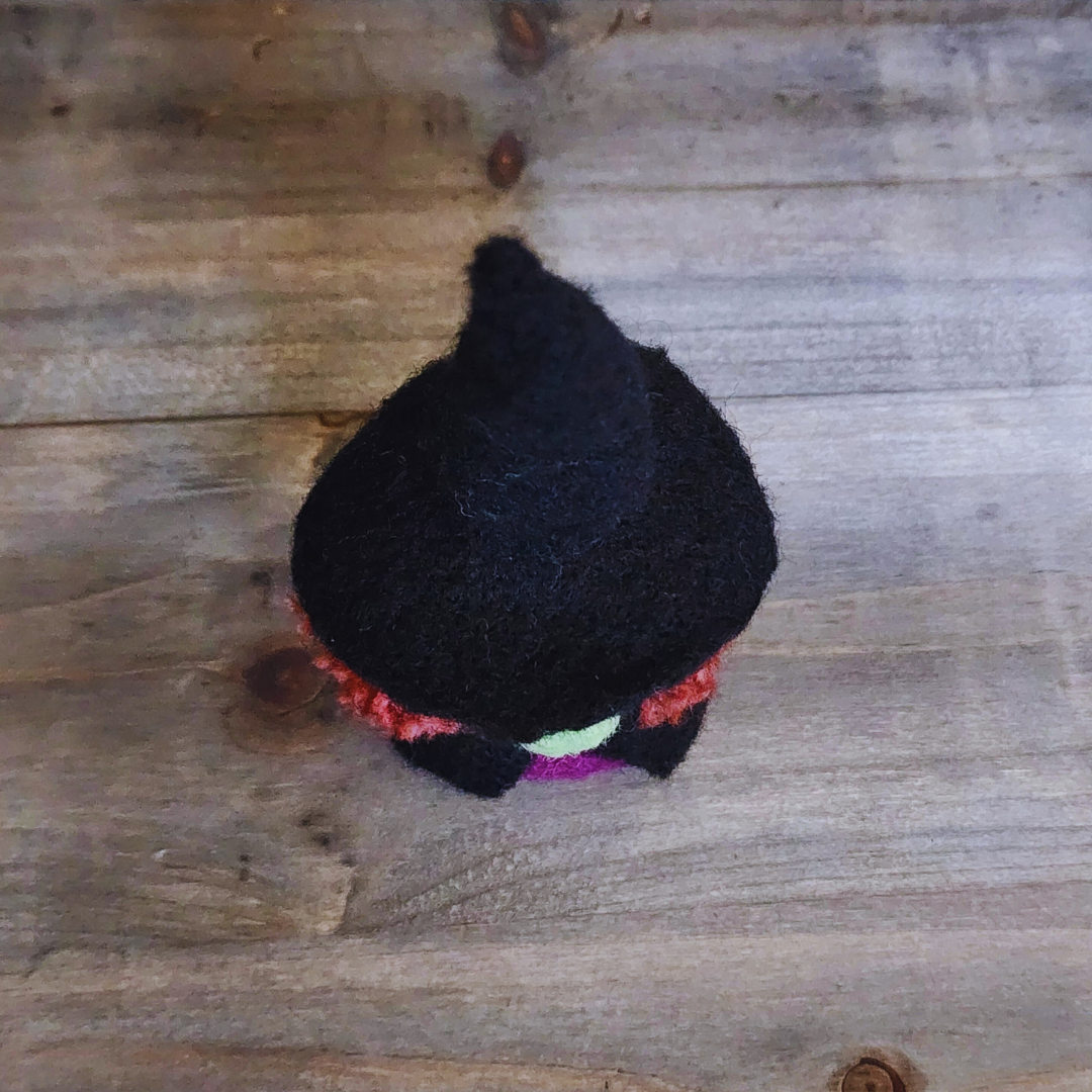 An overhead view of a handmade needle-felted witch figurine, showing the top of its black pointy hat with hints of a green face and orange-red hair peeking out, on a wooden background.