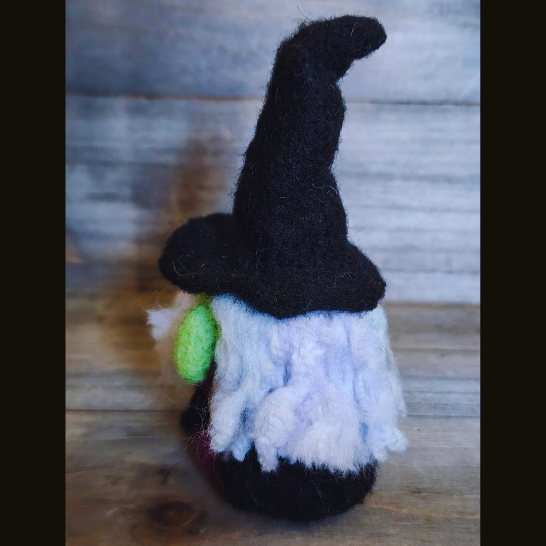 A handmade needle-felted witch figurine with a black pointy hat, a bright green face, a purple body, and fluffy white hair, viewed from a side angle against a wooden background.