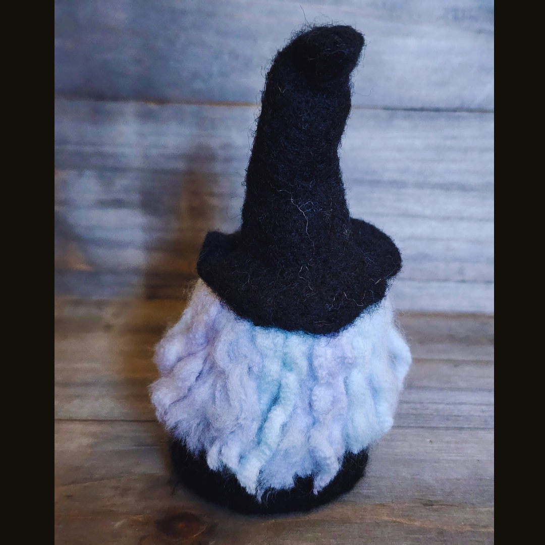 A handmade needle-felted witch figurine with a black pointy hat, a light green face, a purple body, and fluffy white hair, viewed from a slightly angled back position against a wooden background.