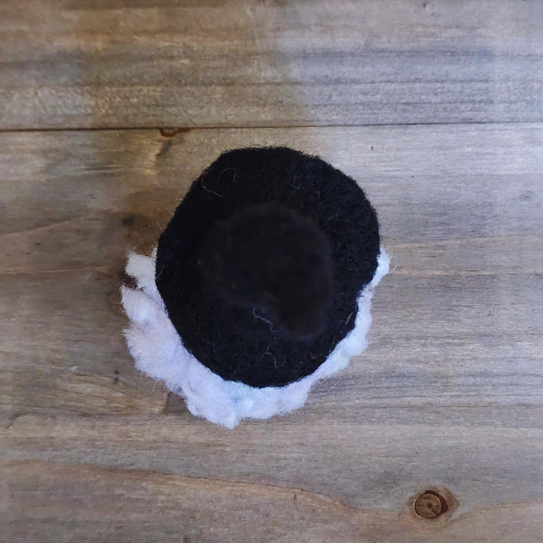 An overhead view of a handmade needle-felted witch figurine with a black pointy hat and fluffy white hair, on a wooden background.