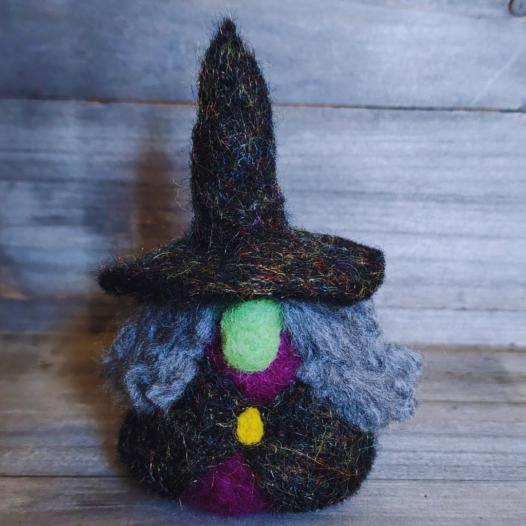 A handmade needle-felted witch figurine with a black sparkly pointy hat, a bright green face, a purple body, a yellow buckle, and fluffy grey hair, standing centered against a wooden background.