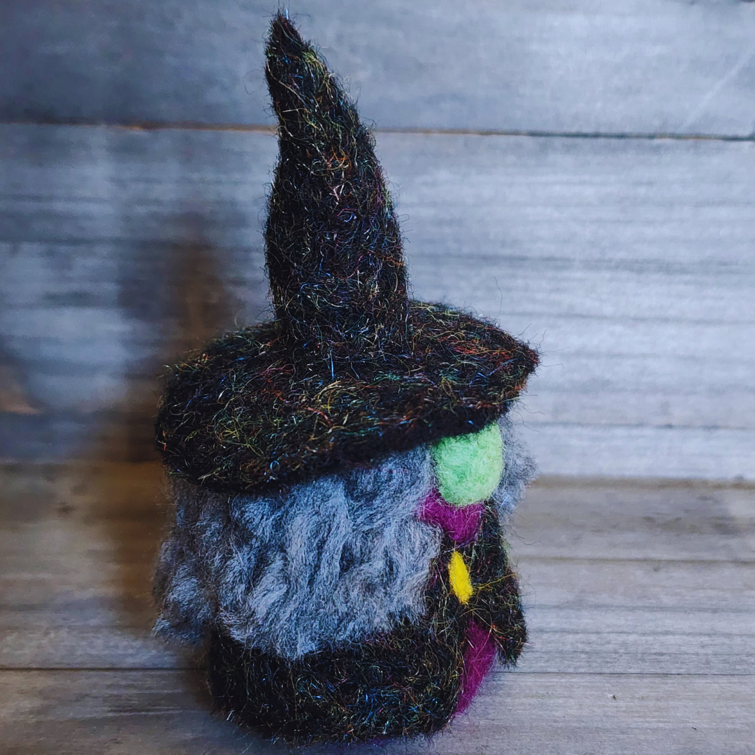 A handmade needle-felted witch figurine with a black sparkly pointy hat, a bright green face, a purple body, a yellow buckle, and fluffy grey hair, viewed from a slightly angled position against a wooden background.