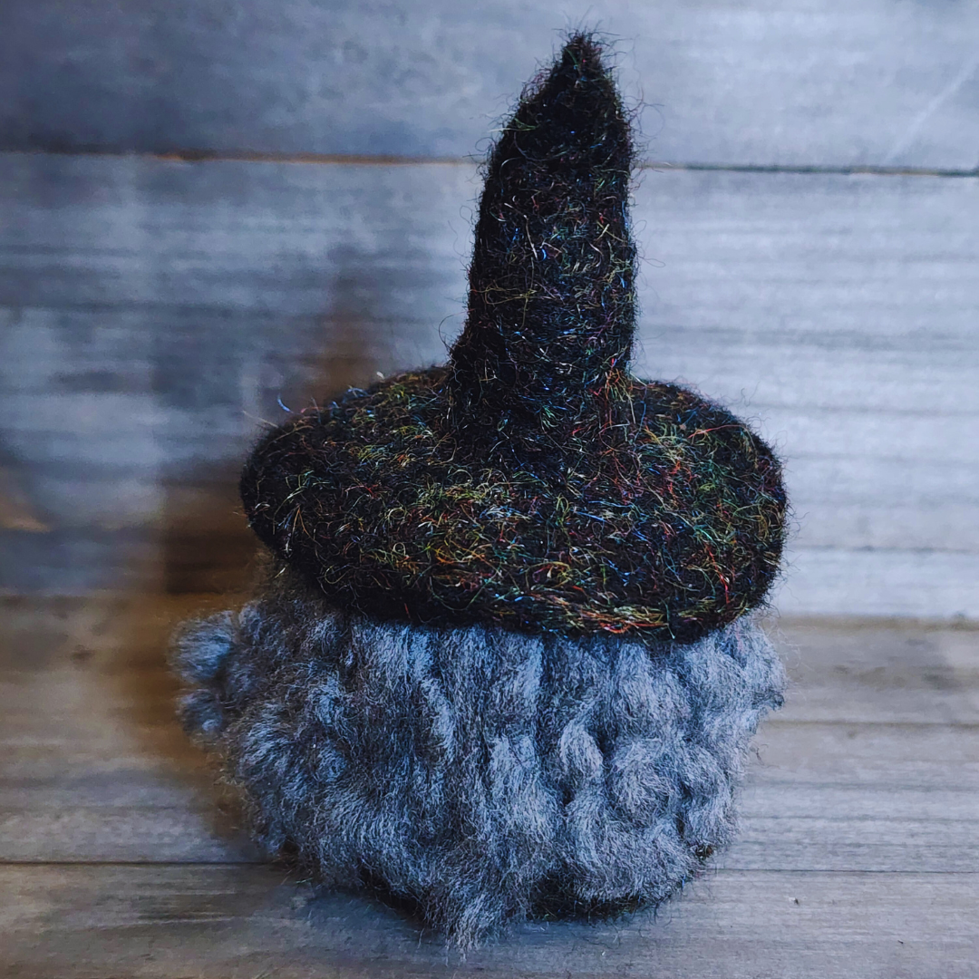 A handmade needle-felted witch figurine with a black sparkly pointy hat and fluffy grey hair, viewed from a side angle against a wooden background.