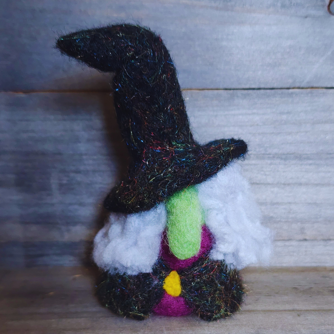 A handmade needle-felted witch figurine with a black sparkly pointy hat, a bright green face, a purple body, a yellow buckle, and fluffy white hair, viewed from a side angle against a wooden background.