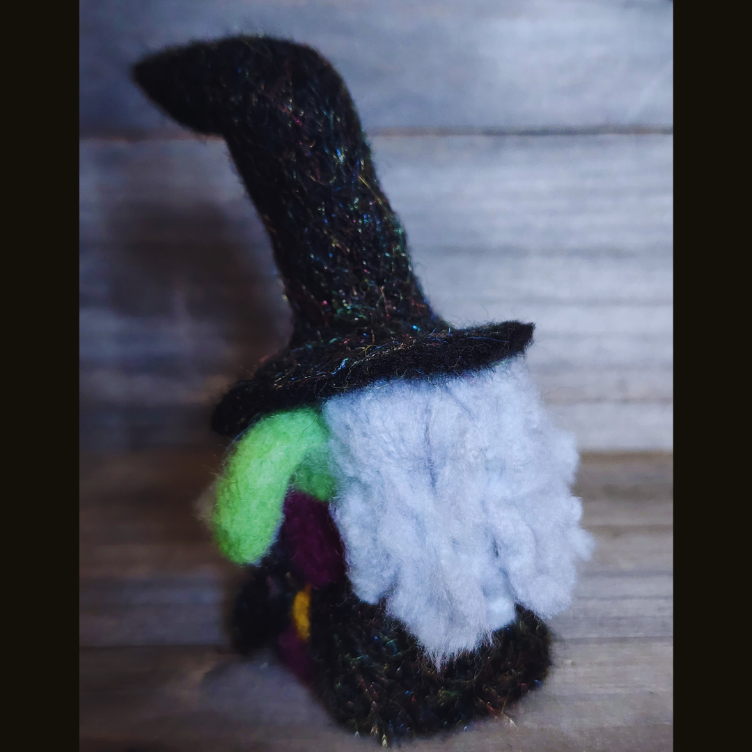  A handmade needle-felted witch figurine with a black sparkly pointy hat, a bright green face, a purple body, a yellow buckle, and fluffy white hair, viewed from a slightly angled position against a wooden background.