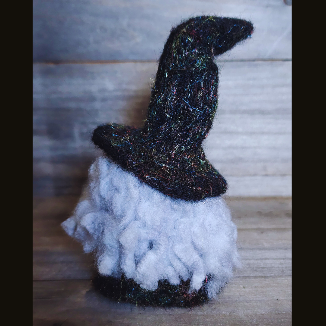 A handmade needle-felted witch figurine with a black sparkly pointy hat and fluffy white hair, viewed from a side angle against a wooden background.
