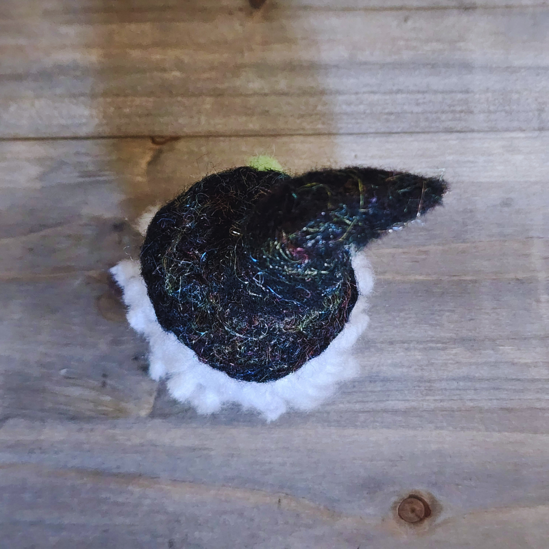 An overhead view of a handmade needle-felted witch figurine with a black sparkly pointy hat and fluffy white hair, on a wooden background.