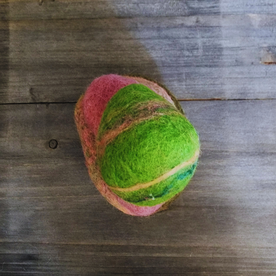 An overhead close-up view of three handmade wet-felted Zen Stones, with a vibrant lime green striped stone stacked on top of a pink-mottled stone, all resting on an olive green stone, on a wooden background.