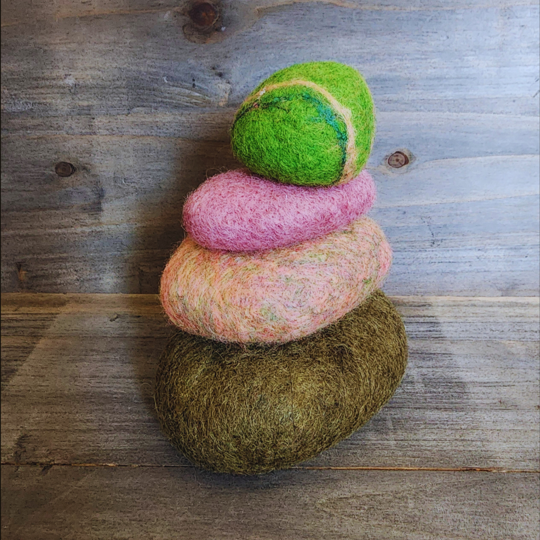 A slightly angled view of four handmade wet-felted Zen Stones stacked, displaying colors of olive green, mottled pink and light green, solid pink, and a vibrant lime green with subtle blue and yellow stripes, on a rustic wooden background.