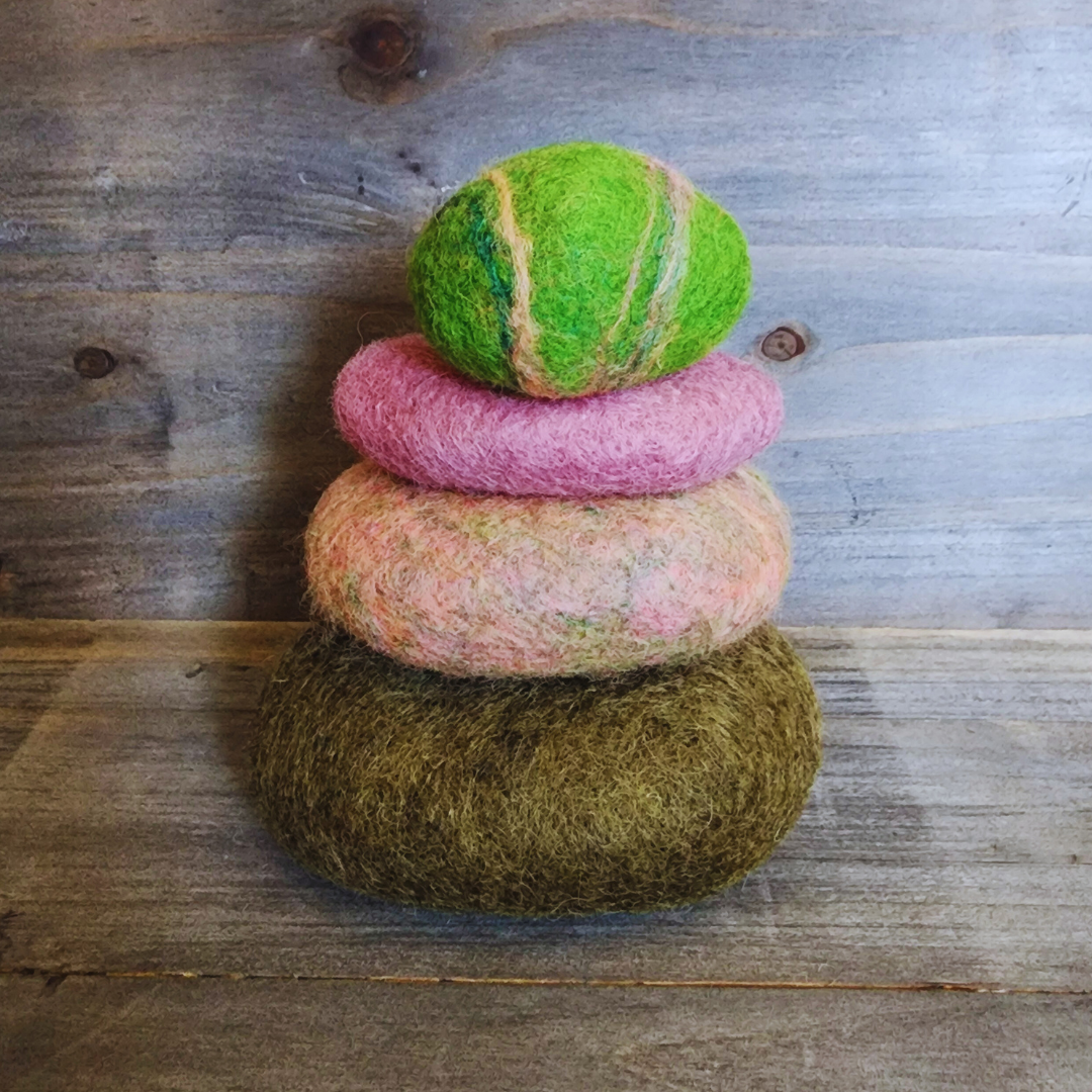 Four handmade wet-felted Zen Stones stacked vertically, from bottom to top: olive green, mottled pink and light green, solid pink, and a vibrant lime green with subtle stripes, on a wooden surface.