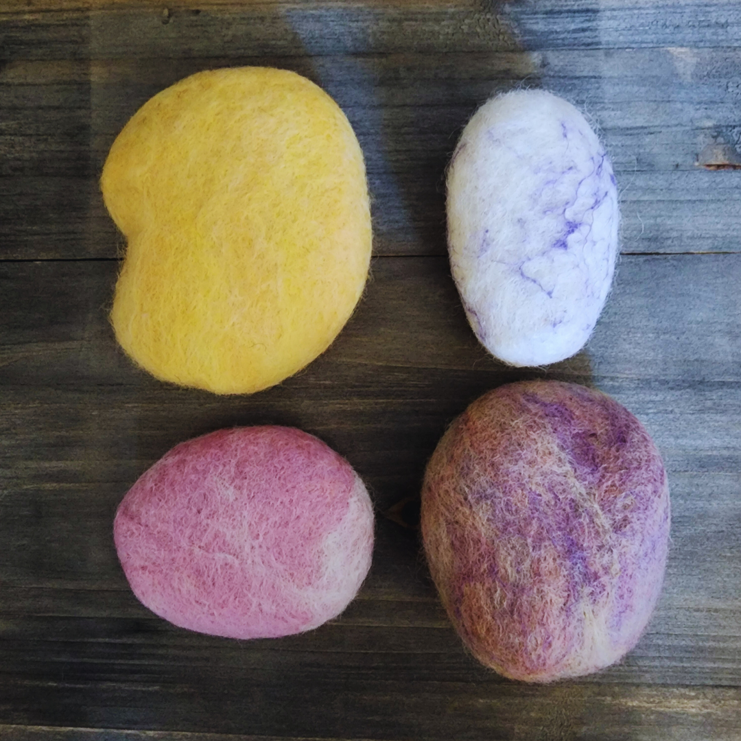 Four handmade wet-felted Zen Stones in an assortment of colors (yellow, light purple/white swirl, pink, and purple mix), arranged unstacked on a wooden background.