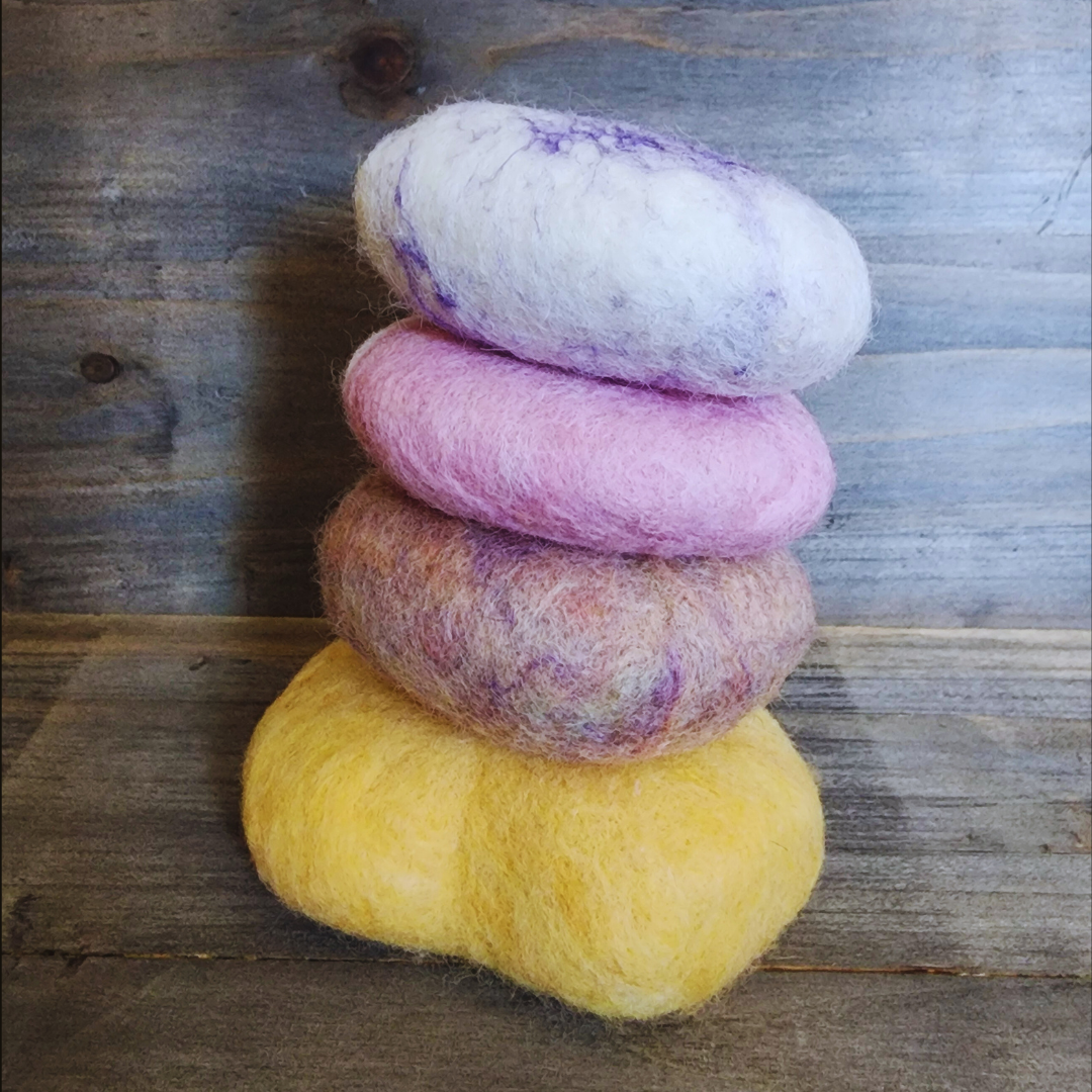 Four handmade wet-felted Zen Stones stacked vertically in an irregular column, featuring colors of yellow, purple mix, pink, and light purple/white swirl, on a wooden background.