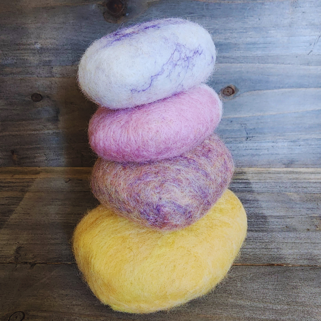 Four handmade wet-felted Zen Stones stacked vertically, from bottom to top: yellow, purple mix, pink, and light purple/white swirl, on a textured wooden background.