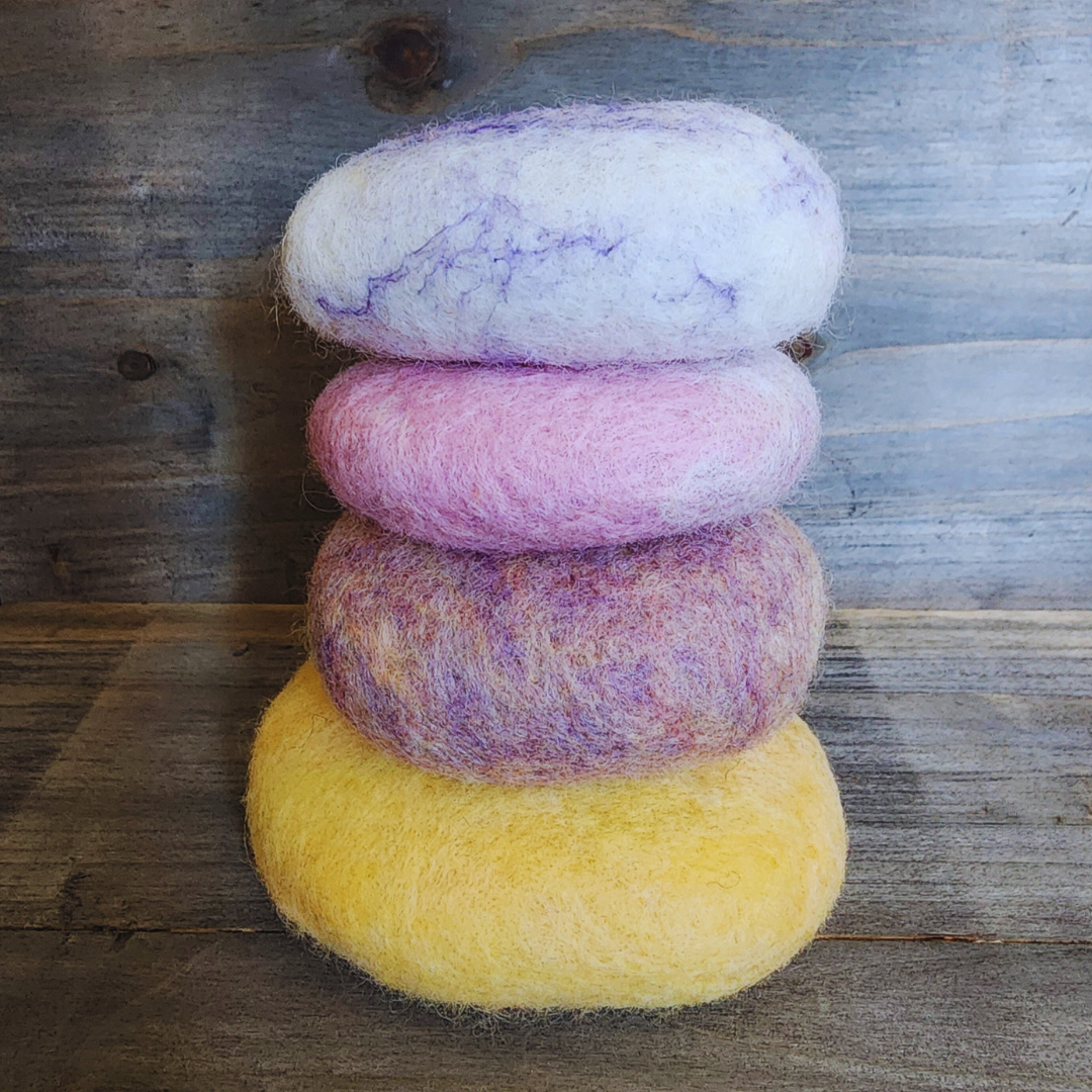 Four handmade wet-felted Zen Stones stacked vertically, from bottom to top: yellow, purple mix, pink, and light purple/white swirl, on a wooden surface.