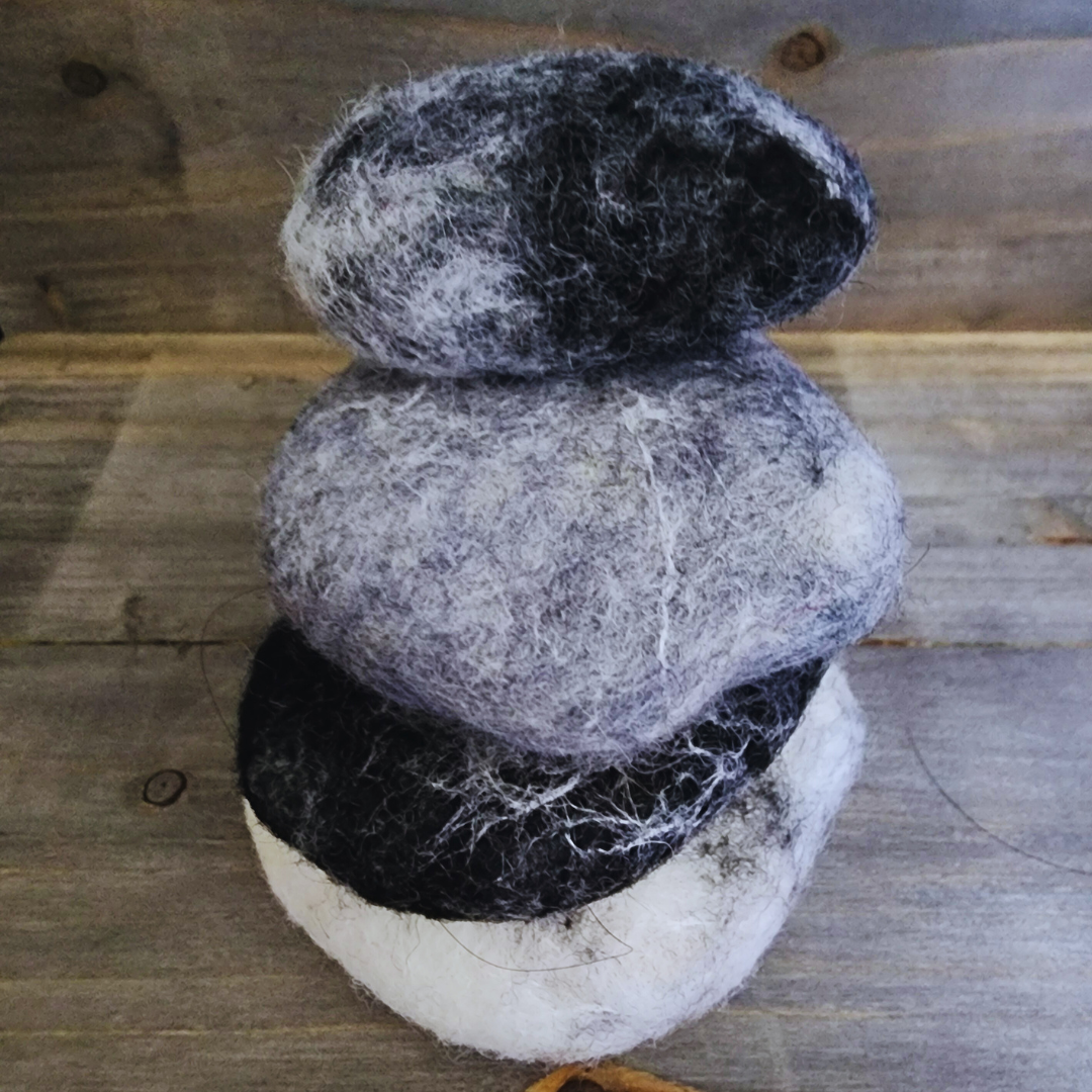 Three handmade wet-felted Zen Stones stacked vertically, featuring colors of dark gray, light gray, and black and white, against a wooden background.