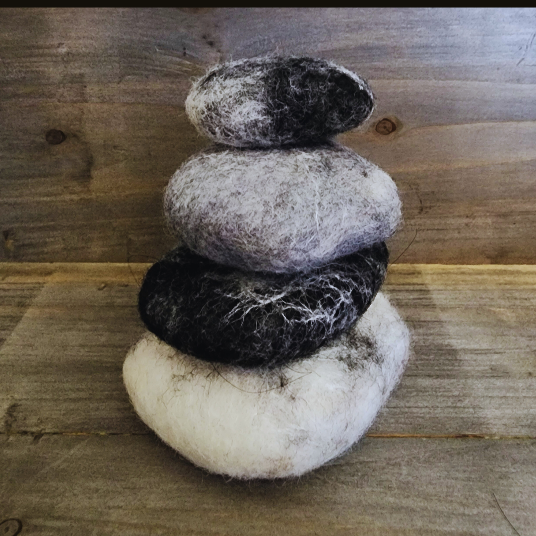 Four handmade wet-felted Zen Stones stacked vertically, showing colors of white, black, light gray, and dark gray, against a textured wooden background.
