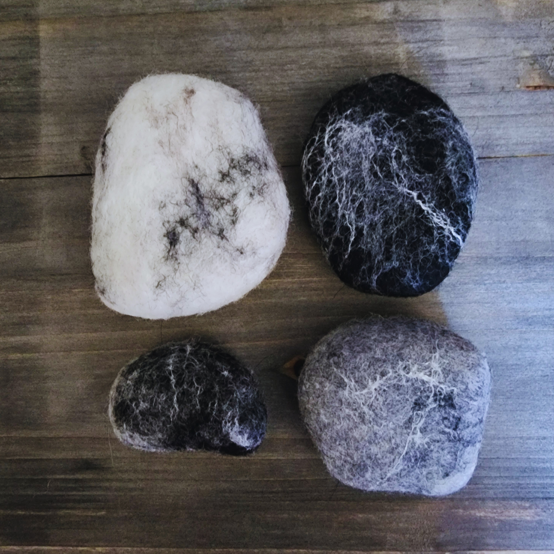 Four handmade wet-felted Zen Stones in various shades of black, gray, and white, arranged unstacked on a wooden surface.