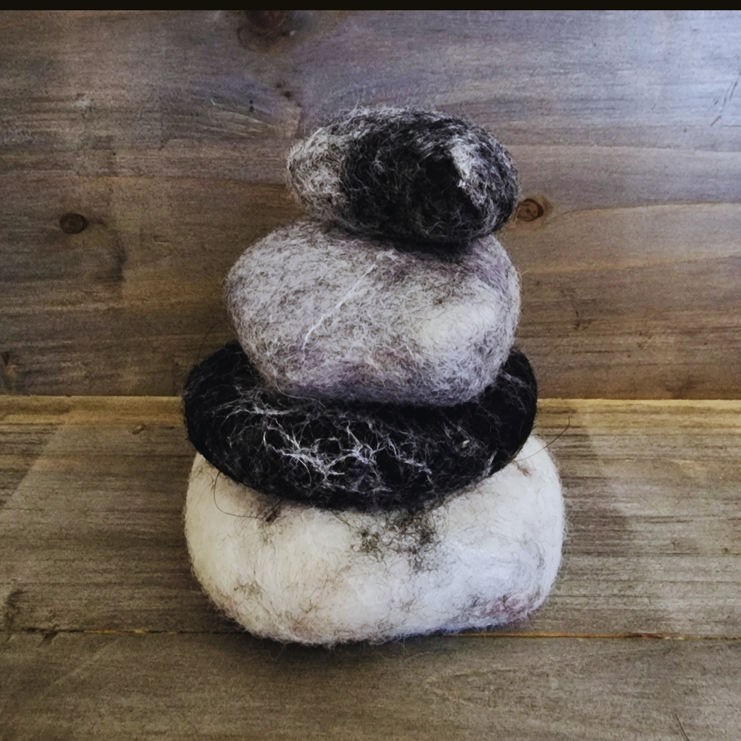 Four handmade wet-felted Zen Stones stacked in a pyramid shape from bottom to top: white, black, light gray, and dark gray, on a wooden surface.