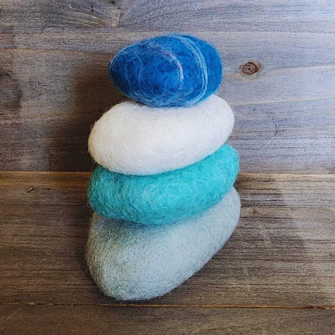 Four handmade wet-felted Zen Stones stacked vertically, featuring shades from bottom to top: light gray, teal, white, and a deep blue with white striations, on a wooden surface.